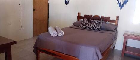 Una cama individual con estructura de madera, una colcha gris y dos toallas enrolladas. Se observan dos adornos en la pared que se asemejan a ojos, una puerta de madera y una pequeña mesa con una silla.