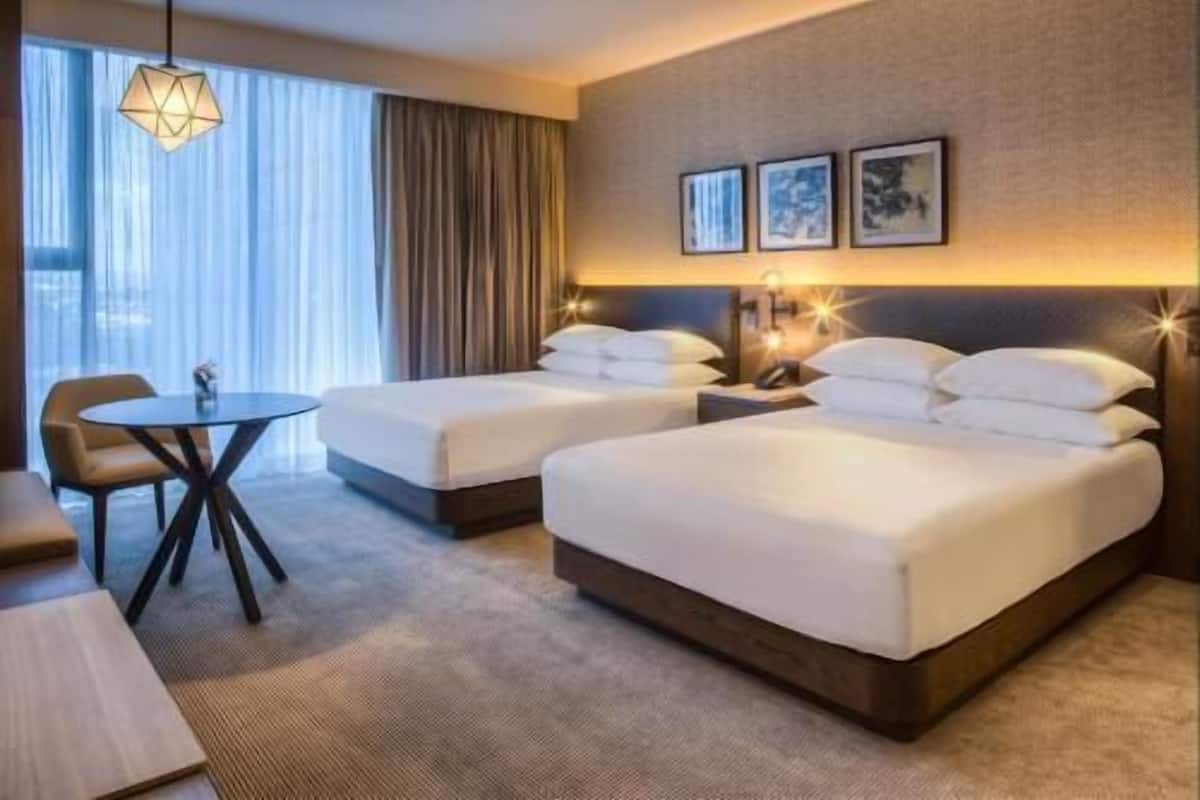 Une chambre d’hôtel avec deux lits, une petite table, une chaise et une grande fenêtre avec des rideaux.