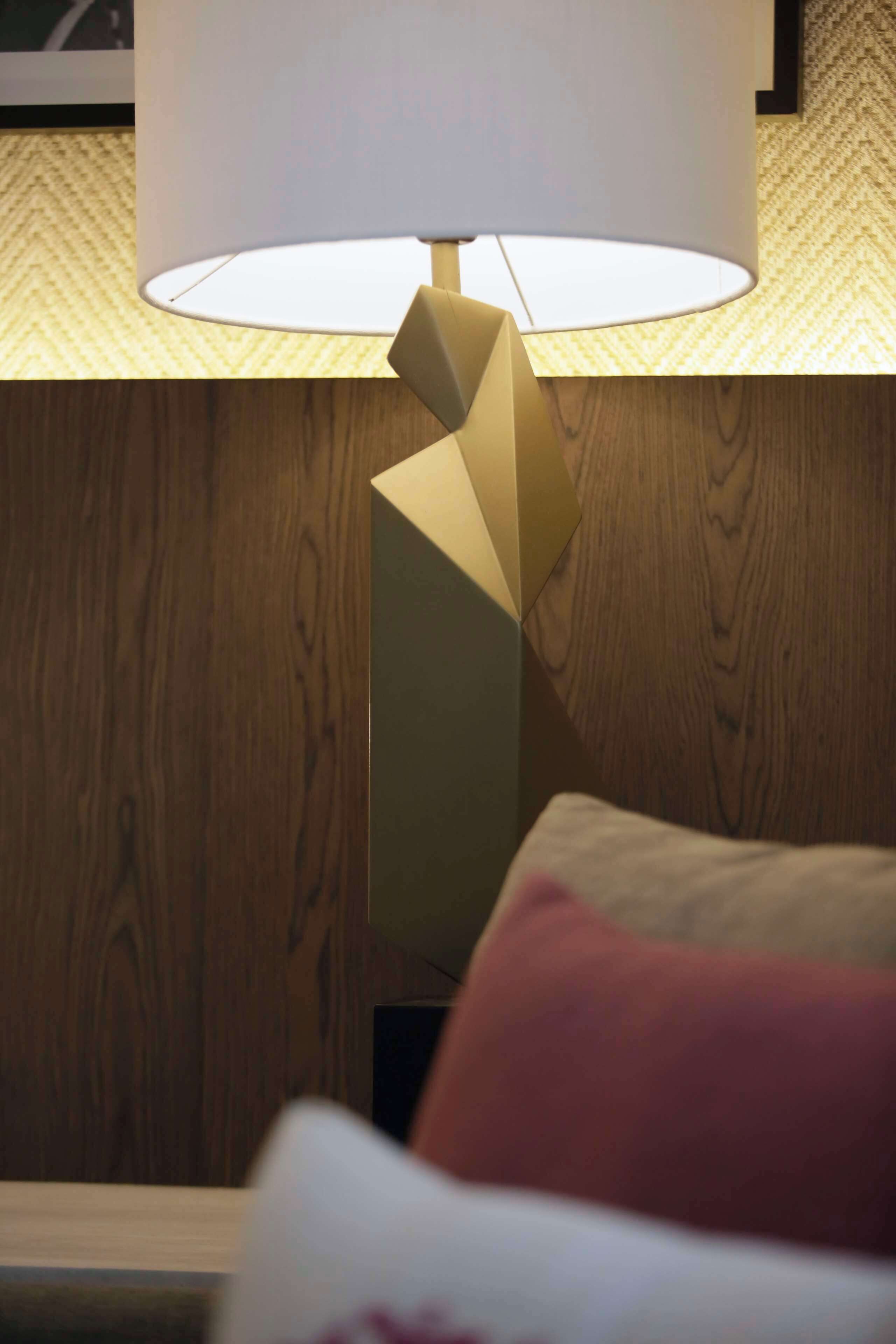 Une lampe moderne dotée d’un pied géométrique et d’un abat-jour blanc, posée sur un plancher en bois, avec un mur à texture en arrière-plan.