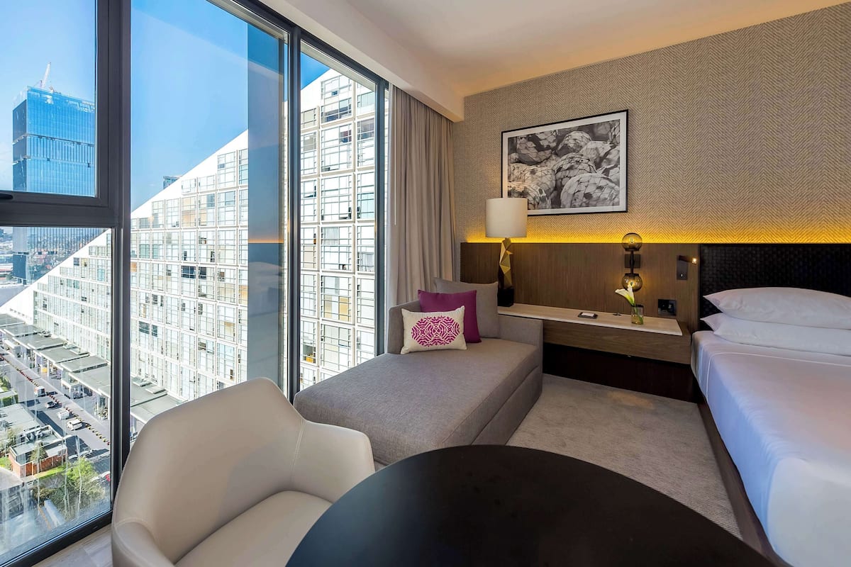 Une chambre d’hôtel avec une grande fenêtre, un canapé, une table de chevet avec une lampe, un lit et un tableau encadré au mur.