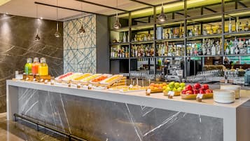 Uma área de bar com balcão de mármore, diversas garrafas, copos e uma exposição de frutas.