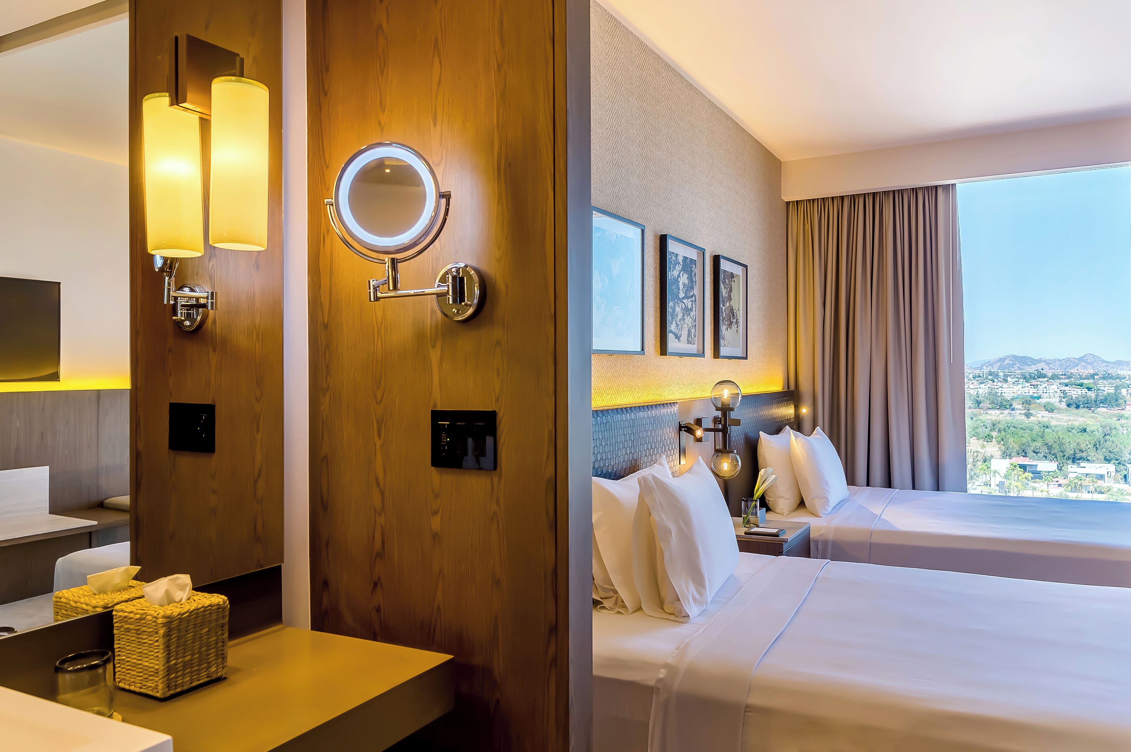 Habitación de hotel con pared de madera, un espejo redondo, cama con ropa blanca, mesita de noche con lámpara y vista a la ciudad a través de amplias ventanas.