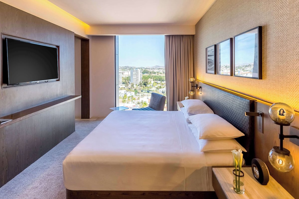 Une chambre d’hôtel dotée d’un grand lit, d’un téléviseur à écran plat fixé au mur, offrant une vue sur la ville grâce à de grandes fenêtres, et comprenant une table de chevet avec une lampe et un vase.
