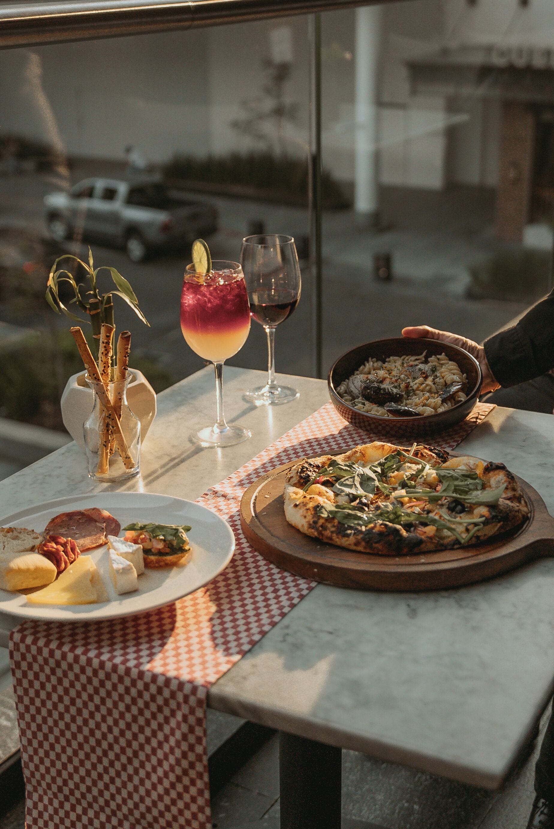 Una mesa con mantel a cuadros, un plato de pasta, una pizza, una copa de vino tinto y una copa de una bebida roja y amarilla.