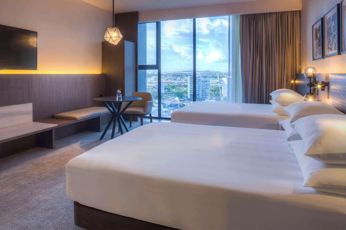 Une chambre d’hôtel avec deux lits, un bureau, un canapé et une grande fenêtre donnant sur la ville.