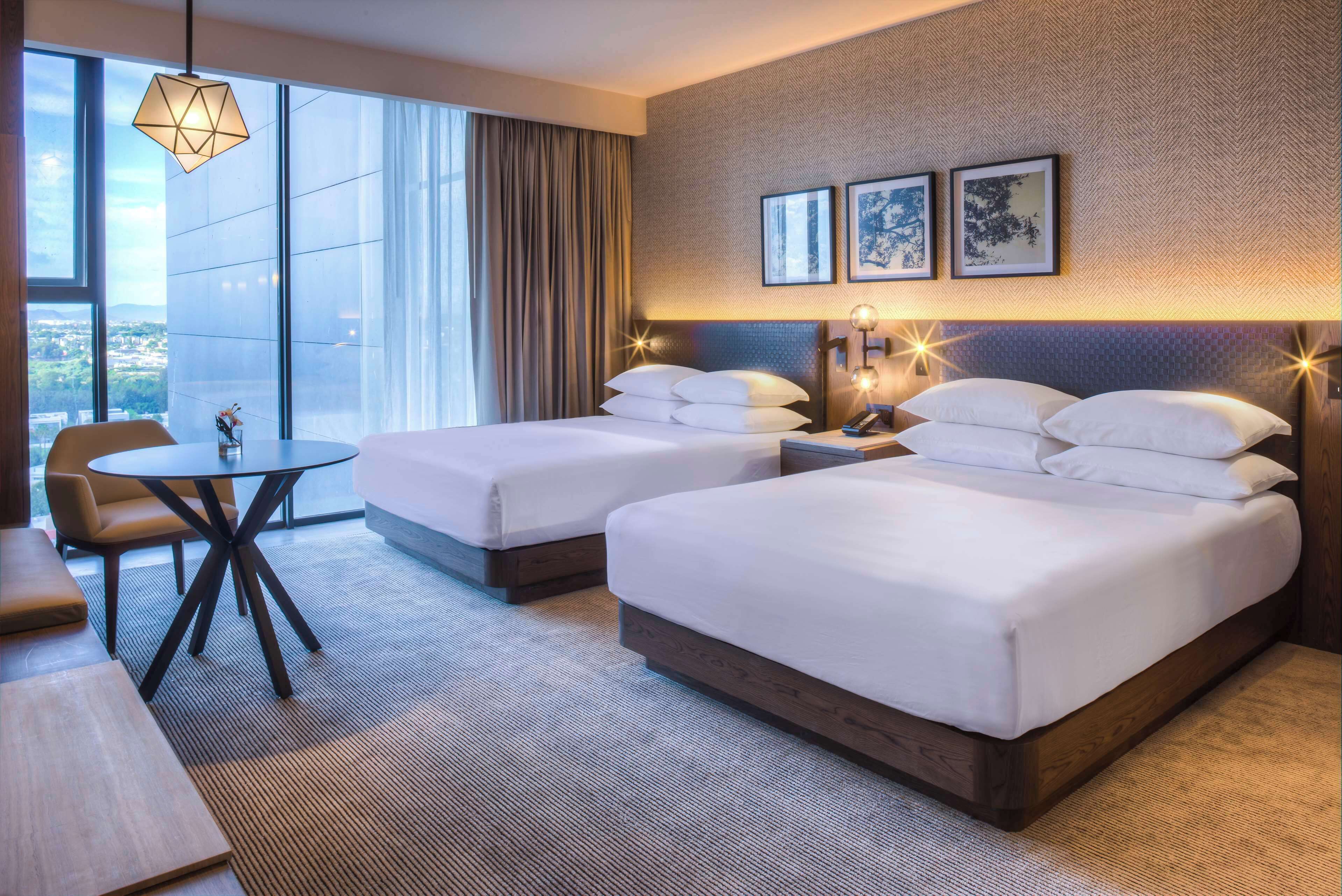 Habitación de hotel con dos camas, una mesa redonda, una silla y amplias ventanas con vistas.
