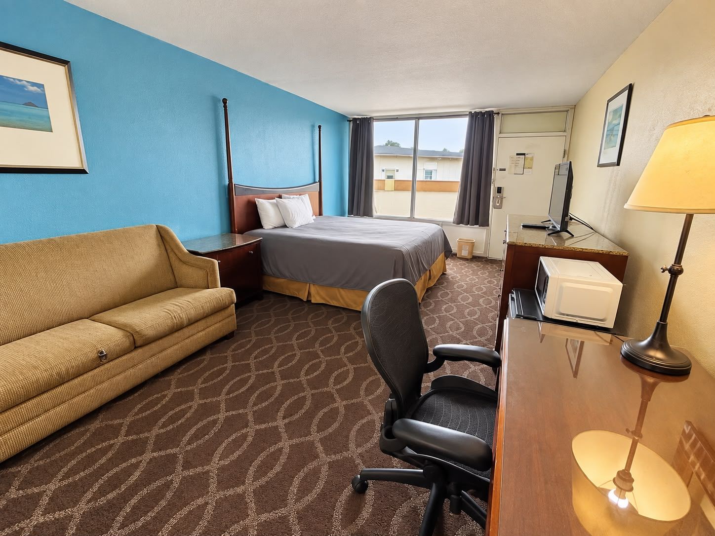 A hotel room with a bed, a sofa, a desk, a chair, a lamp, and a window with curtains.