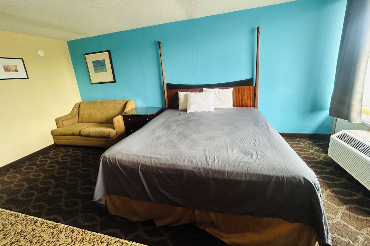 A hotel room with a bed, a chair, a picture on the wall, and an air conditioning unit.