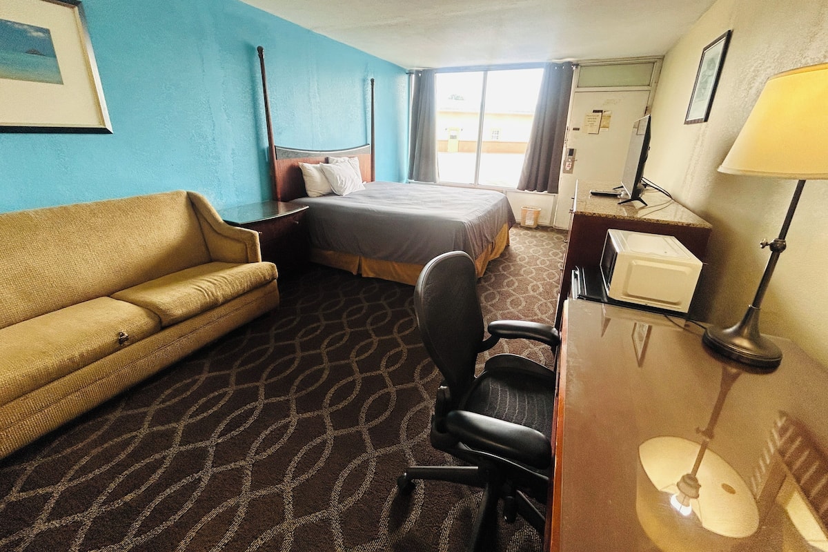 A hotel room with a bed, a desk with a lamp, a chair, a couch, and a window.