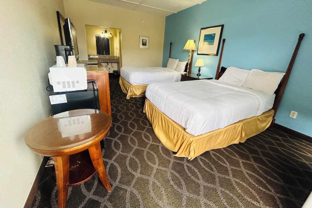 A hotel room with two beds, a desk, a chair, a mirror, a lamp, and a picture on the wall.