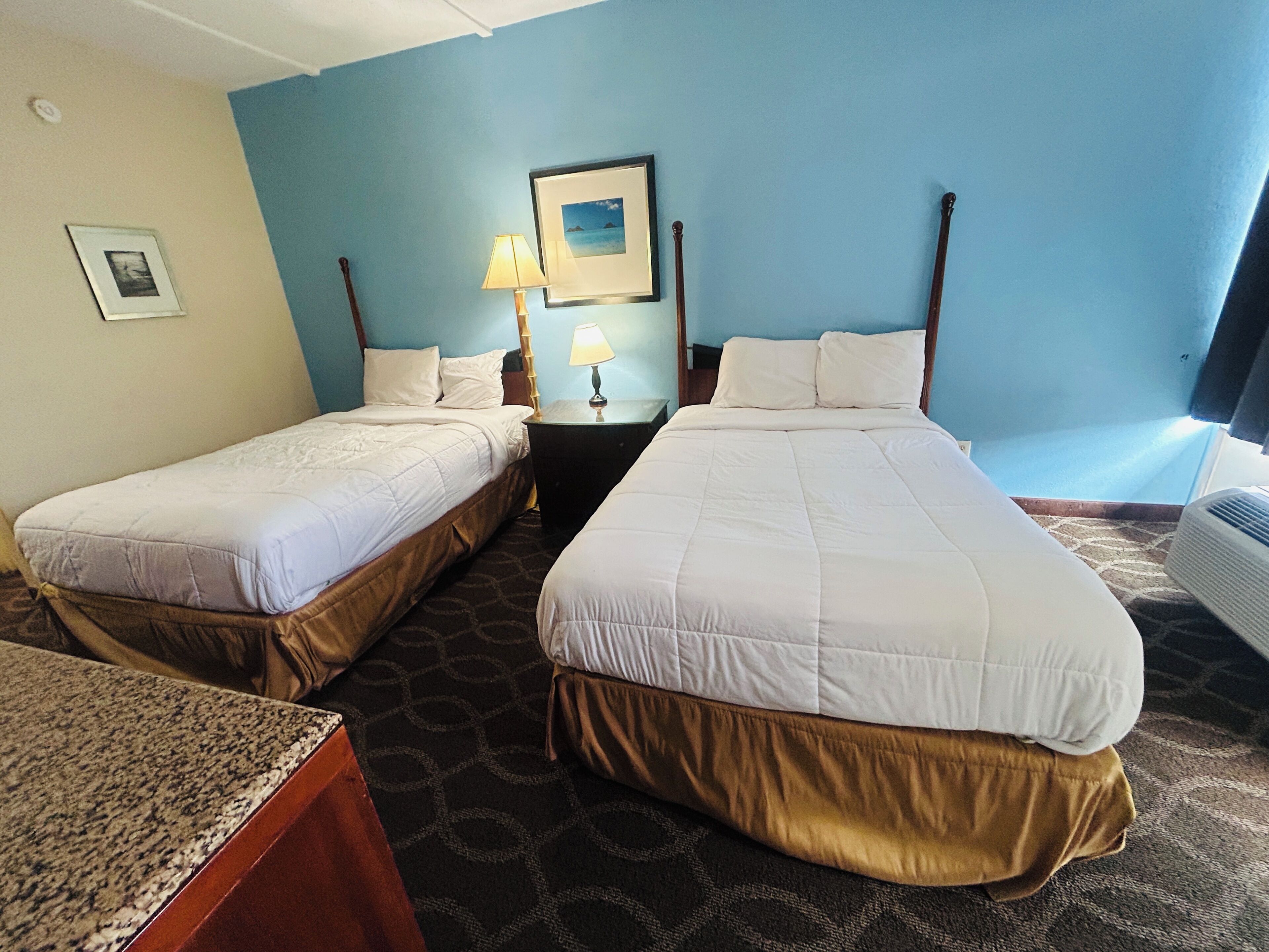Habitación de hotel con dos camas individuales, una mesita de noche con lámpara, un cuadro enmarcado en la pared y una ventana con cortinas.