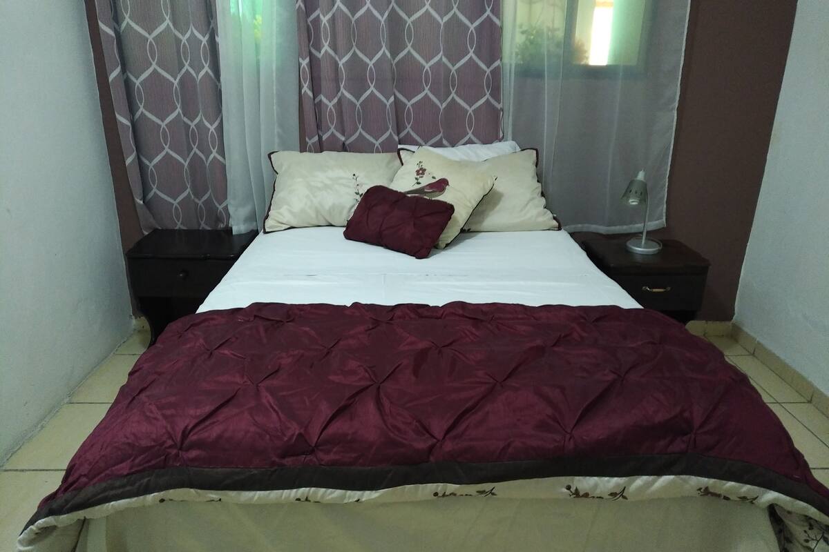 A neatly made bed with a red and beige bedspread, two pillows with floral designs, and a bedside table with a lamp.