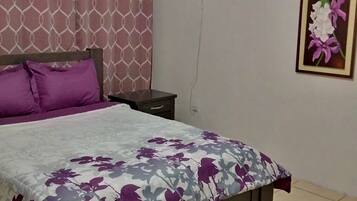 A bedroom with a bed, a nightstand, a wall-mounted fan, and a floral painting.