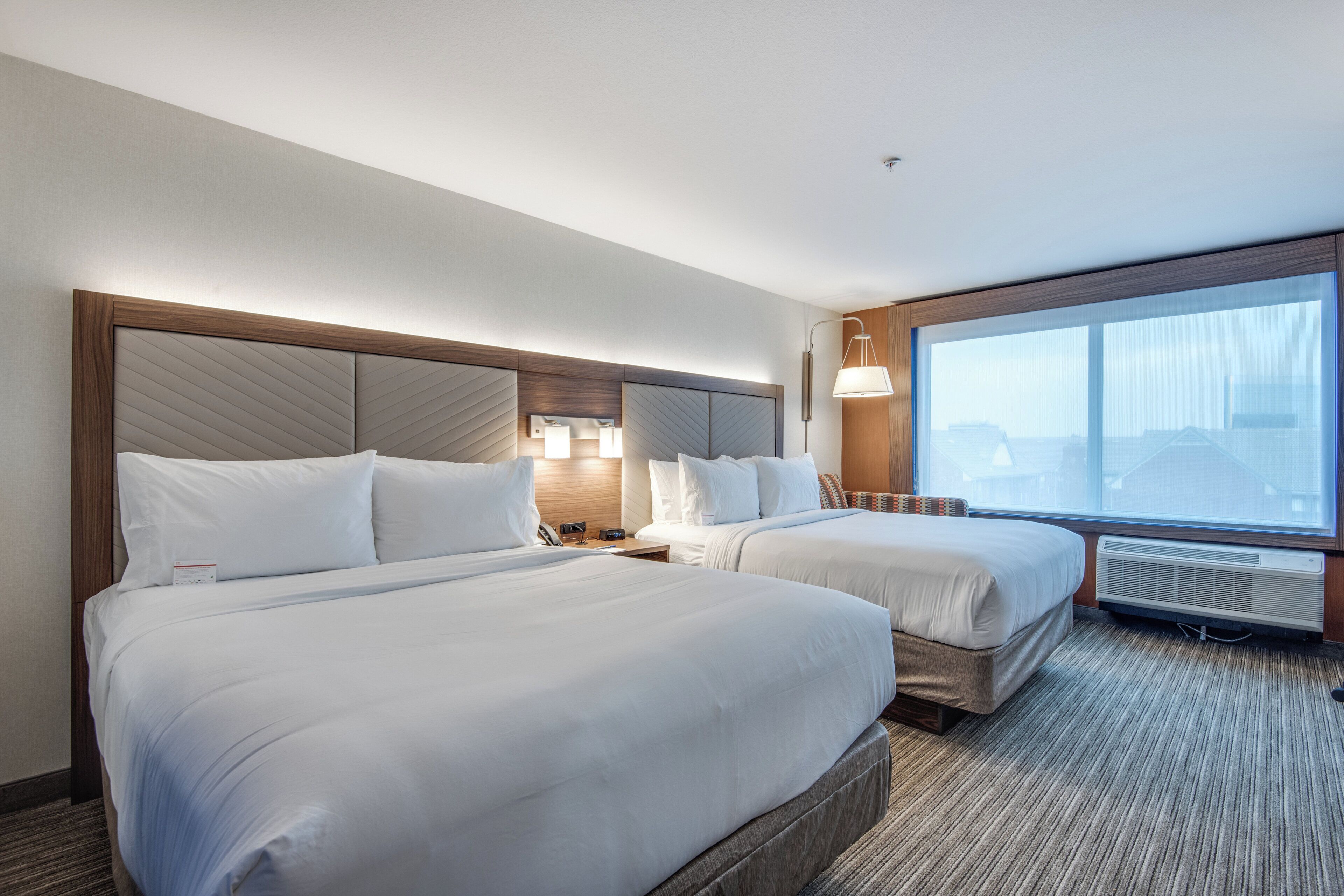 Habitación de hotel con dos camas, un ventanal grande, un calentador de pared y una mesita de noche con lámpara.