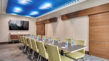 Un salón de conferencias con una larga mesa, sillas, botellas de agua y un televisor en la pared.