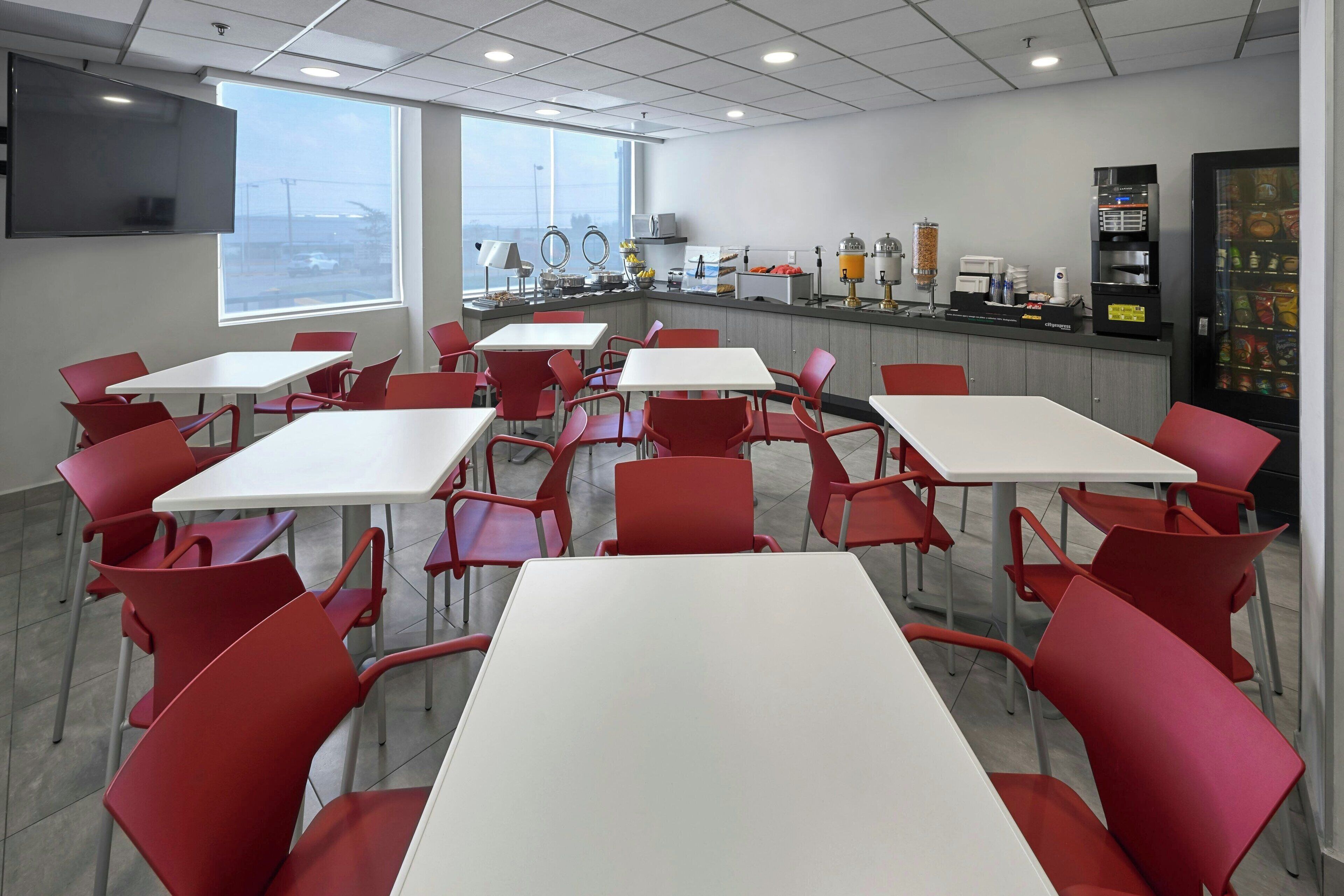 Un área de comedor moderna con sillas rojas y mesas blancas, una encimera de cocina con electrodomésticos y una máquina expendedora.