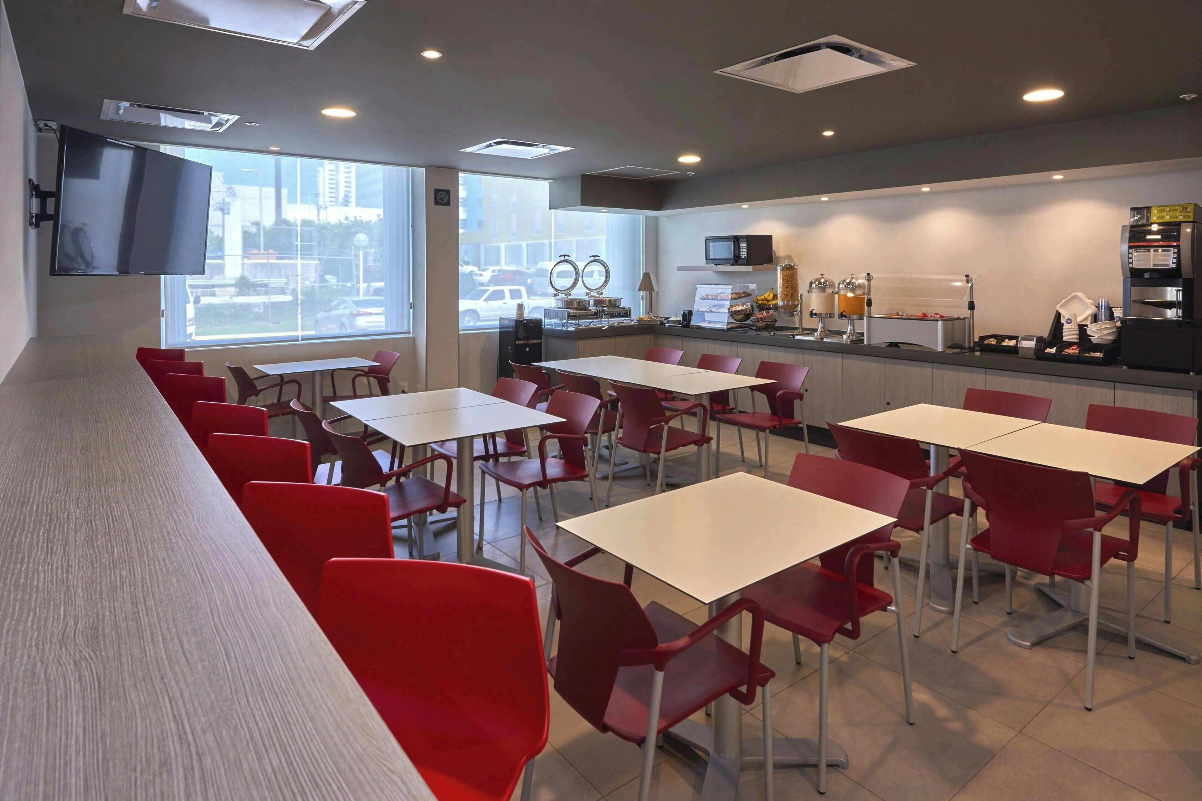 Un espace repas moderne avec des chaises rouges et des tables blanches, un téléviseur à écran plat fixé au mur, et un comptoir de cuisine équipé d’un four à micro-ondes et d’une machine à café.