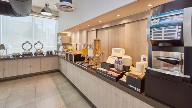 Un área de desayuno de hotel con una larga barra, diversas opciones de comida, una máquina de café y un dispensador de bebidas de autoservicio.