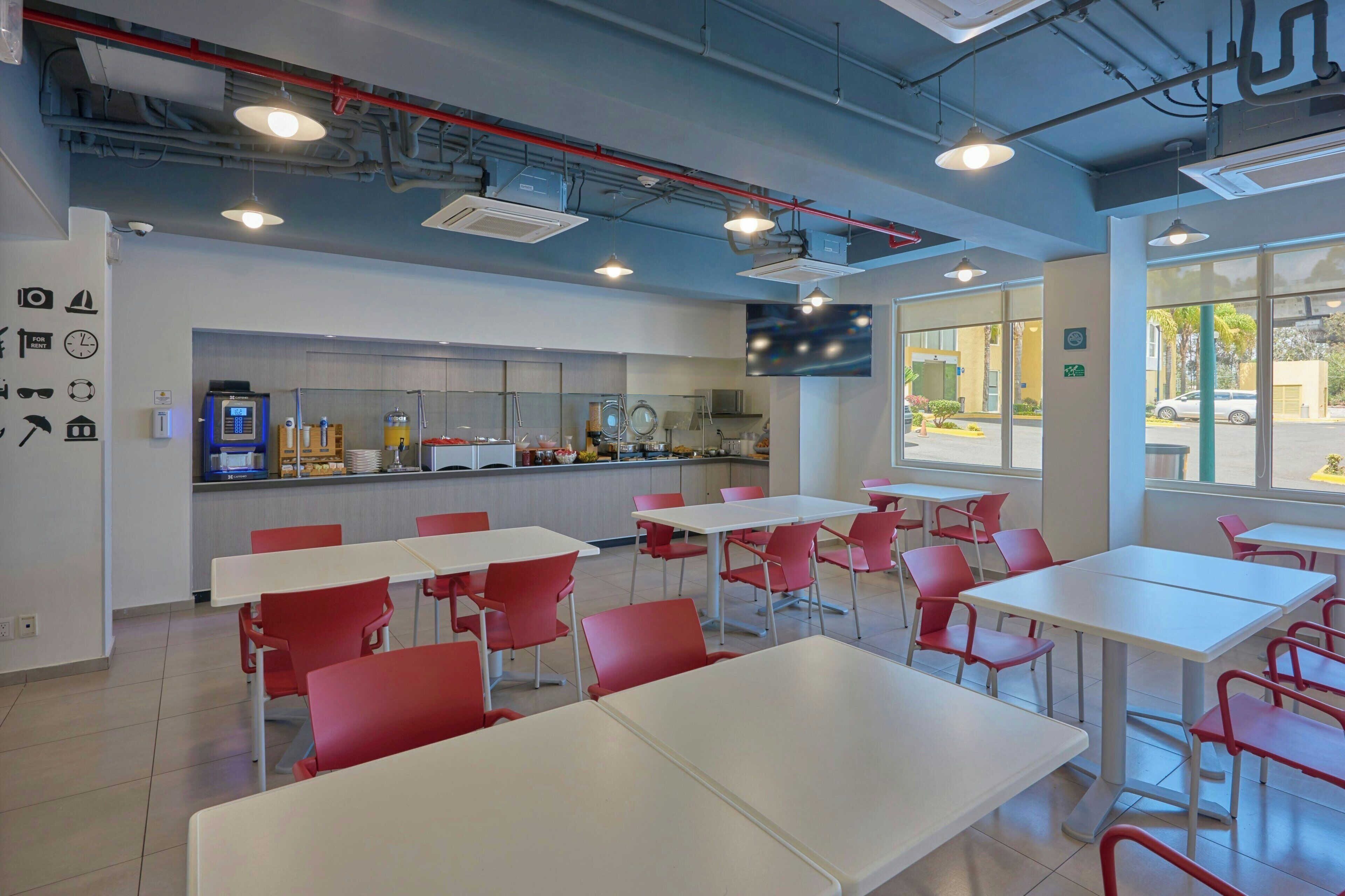 A modern dining area with white tables and red chairs, a kitchen in the background, and large windows.