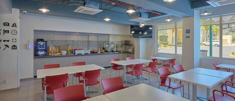 A modern dining area with white tables and red chairs, a kitchen in the background, and large windows.