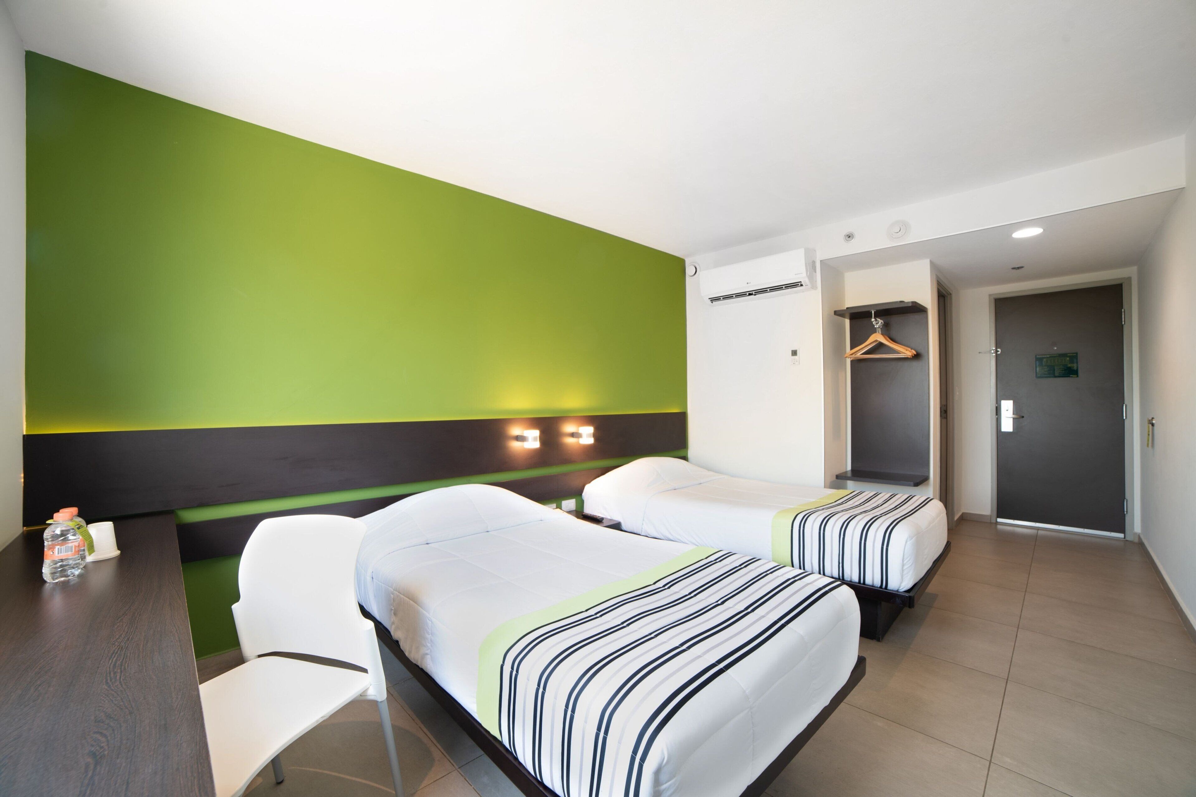 A hotel room with two beds, a desk, and a chair. There is a coat rack and a door to the right.