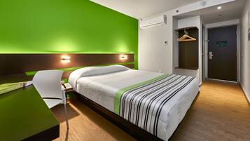 A modern hotel room with a green accent wall, a bed with striped bedding, a desk with a chair, and a door with a hanging coat rack.