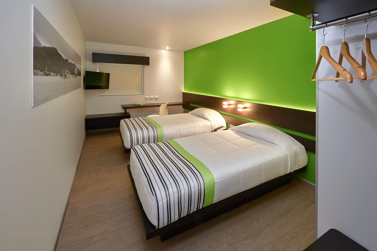 Una habitación de hotel moderna con dos camas, un muro de acento verde, un televisor montado y un perchero de madera.