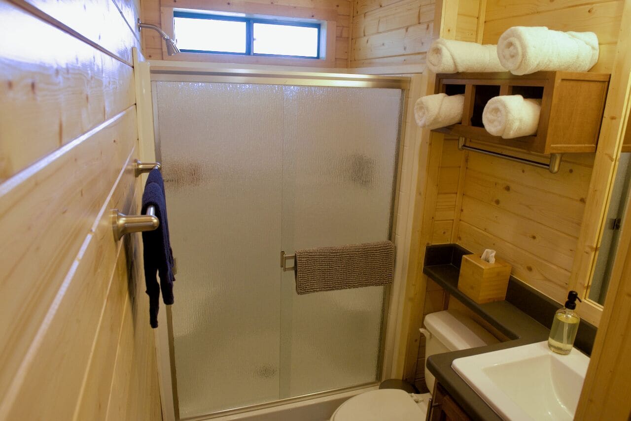 A wooden cabin bathroom with a glass shower door, a towel rack, a sink, and a window above the shower.