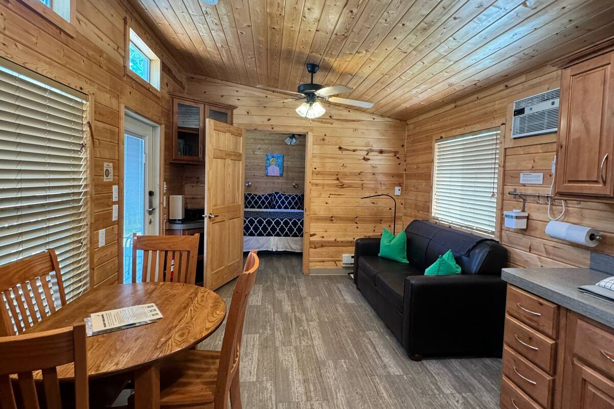 A cabin interior with a wooden dining table, chairs, a black sofa, and a ceiling fan.
