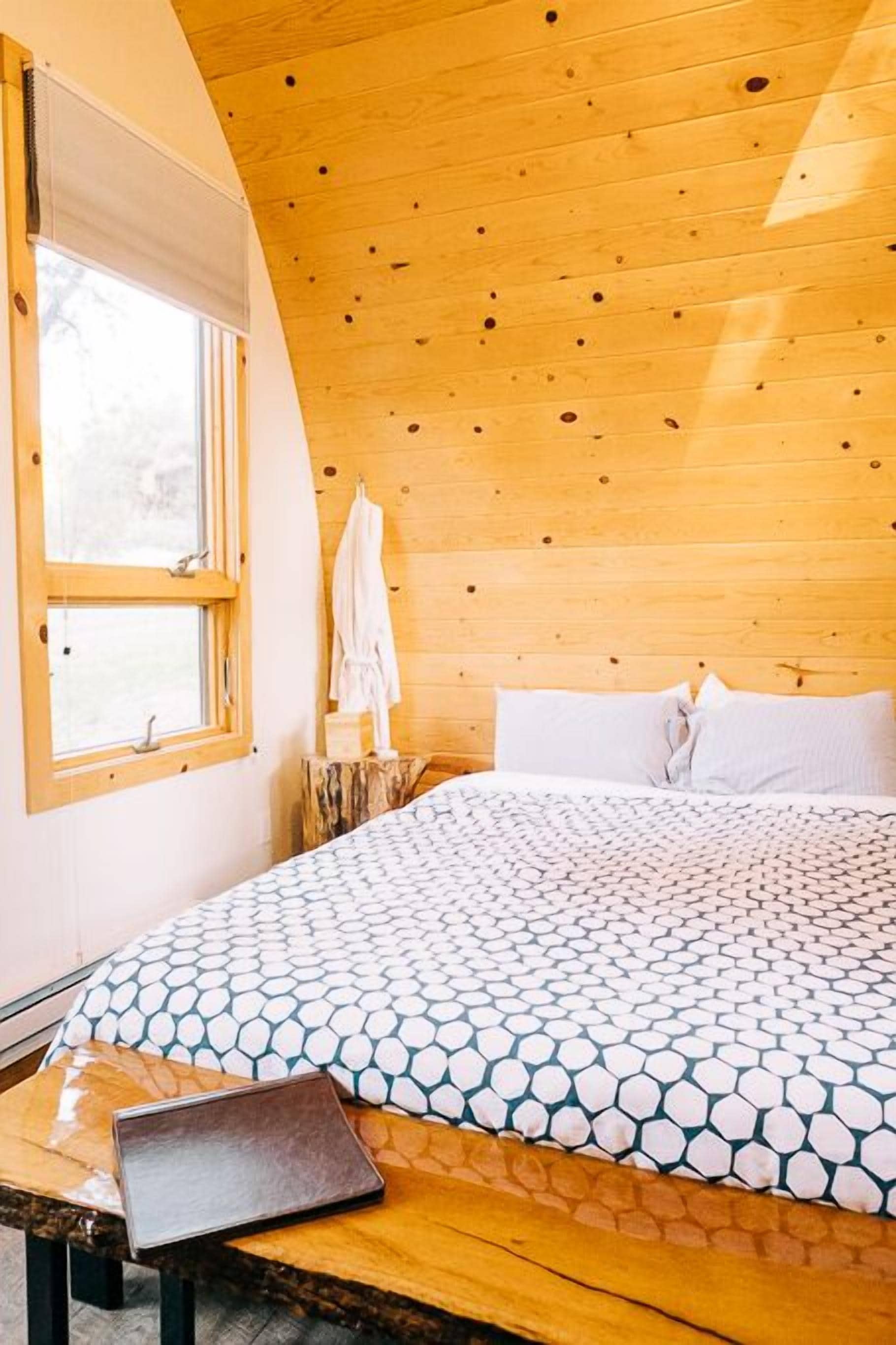 A bedroom with a wooden ceiling, a bed with patterned bedding, a window with a view, and a laptop on a wooden table.