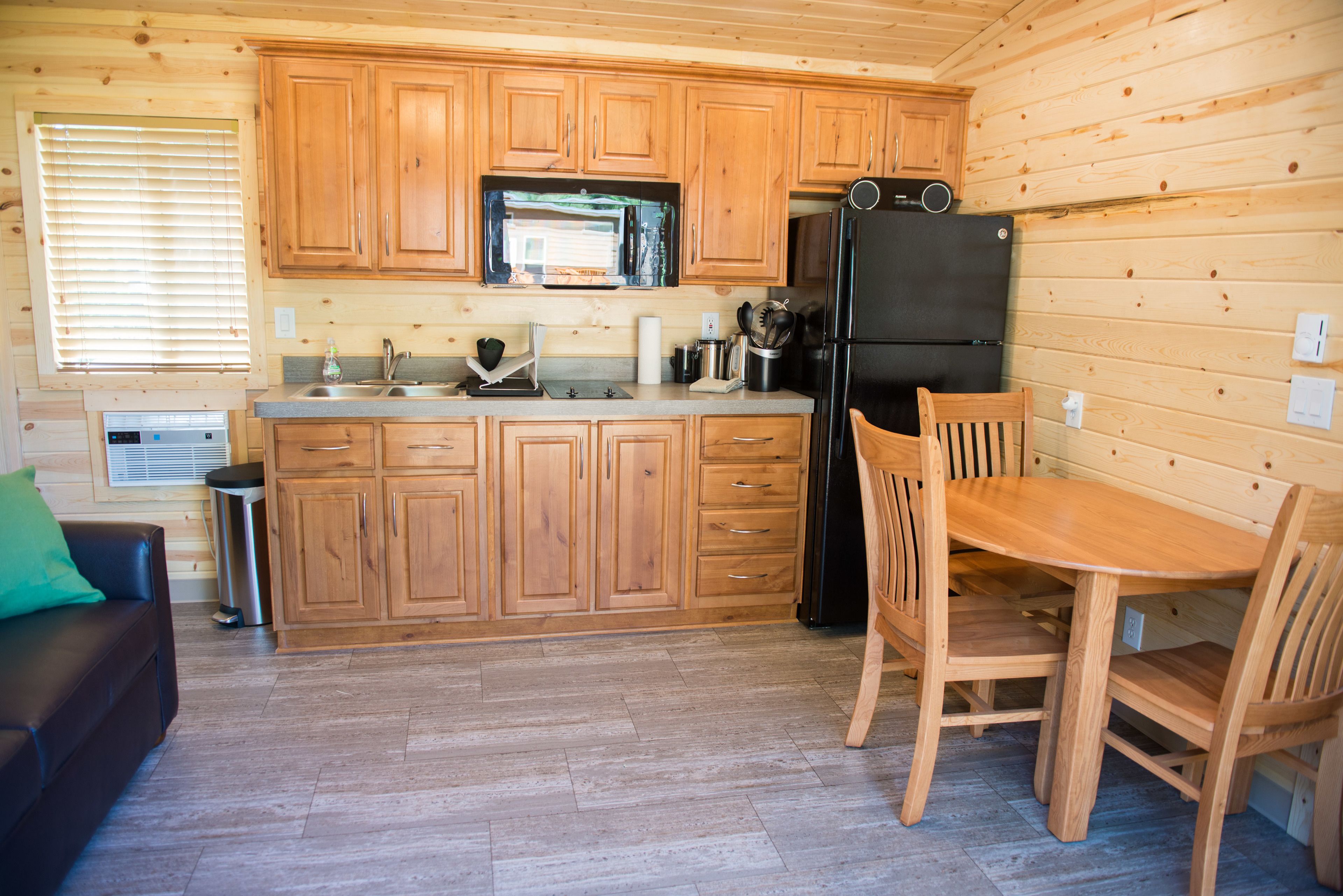 A wooden cabin interior with a kitchen featuring wooden cabinets, a black refrigerator, a microwave, and a dining table with chairs.