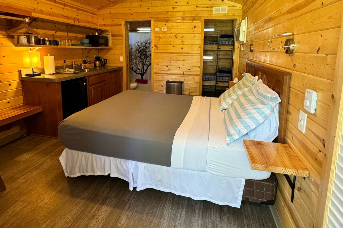 A wooden cabin with a bed, a ceiling fan, a kitchen area, and a bathroom.