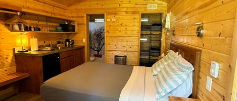 A wooden cabin with a bed, a ceiling fan, a kitchen area, and a bathroom.