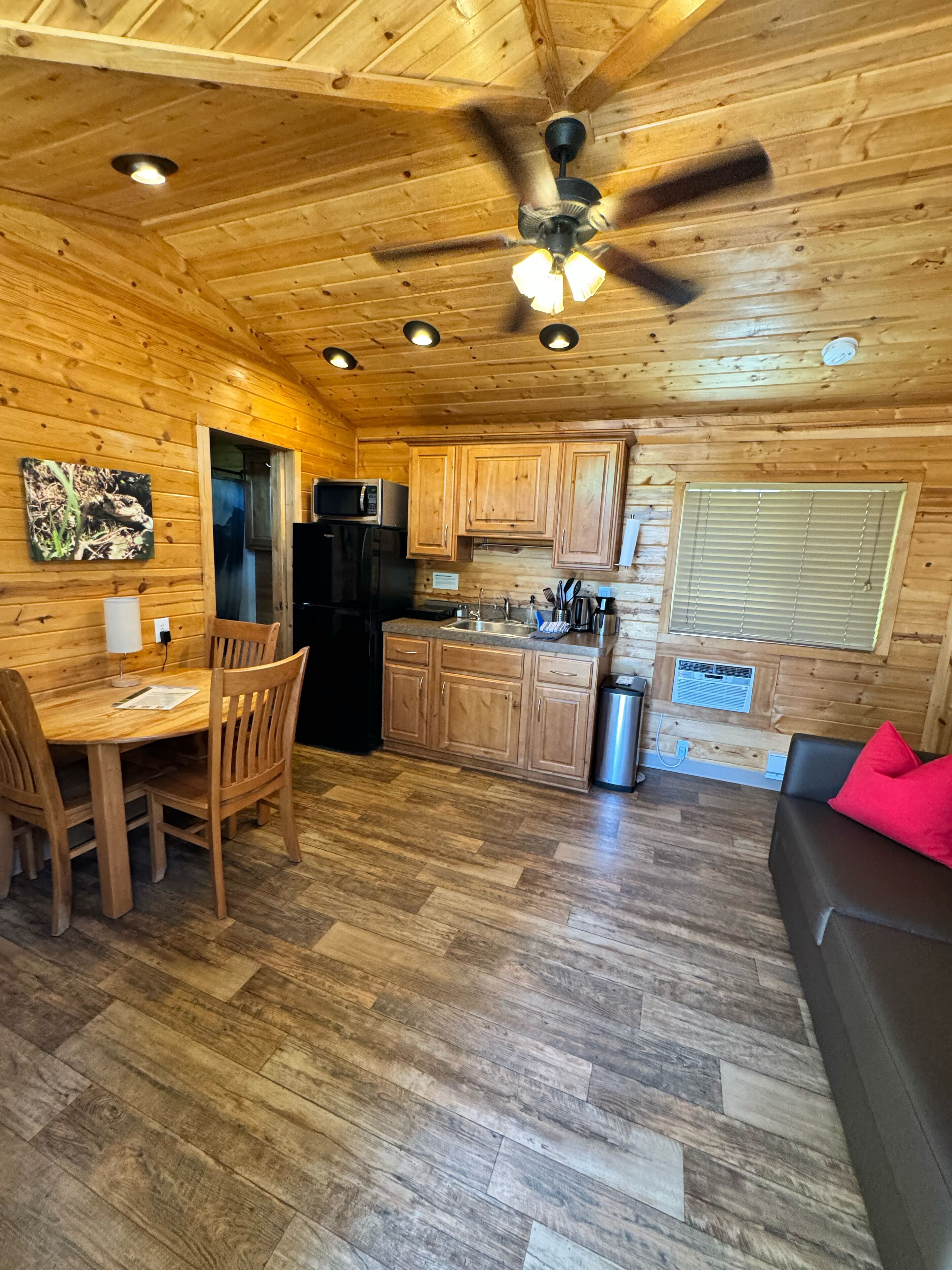 A wooden cabin interior with a dining area, kitchen, and a ceiling fan.