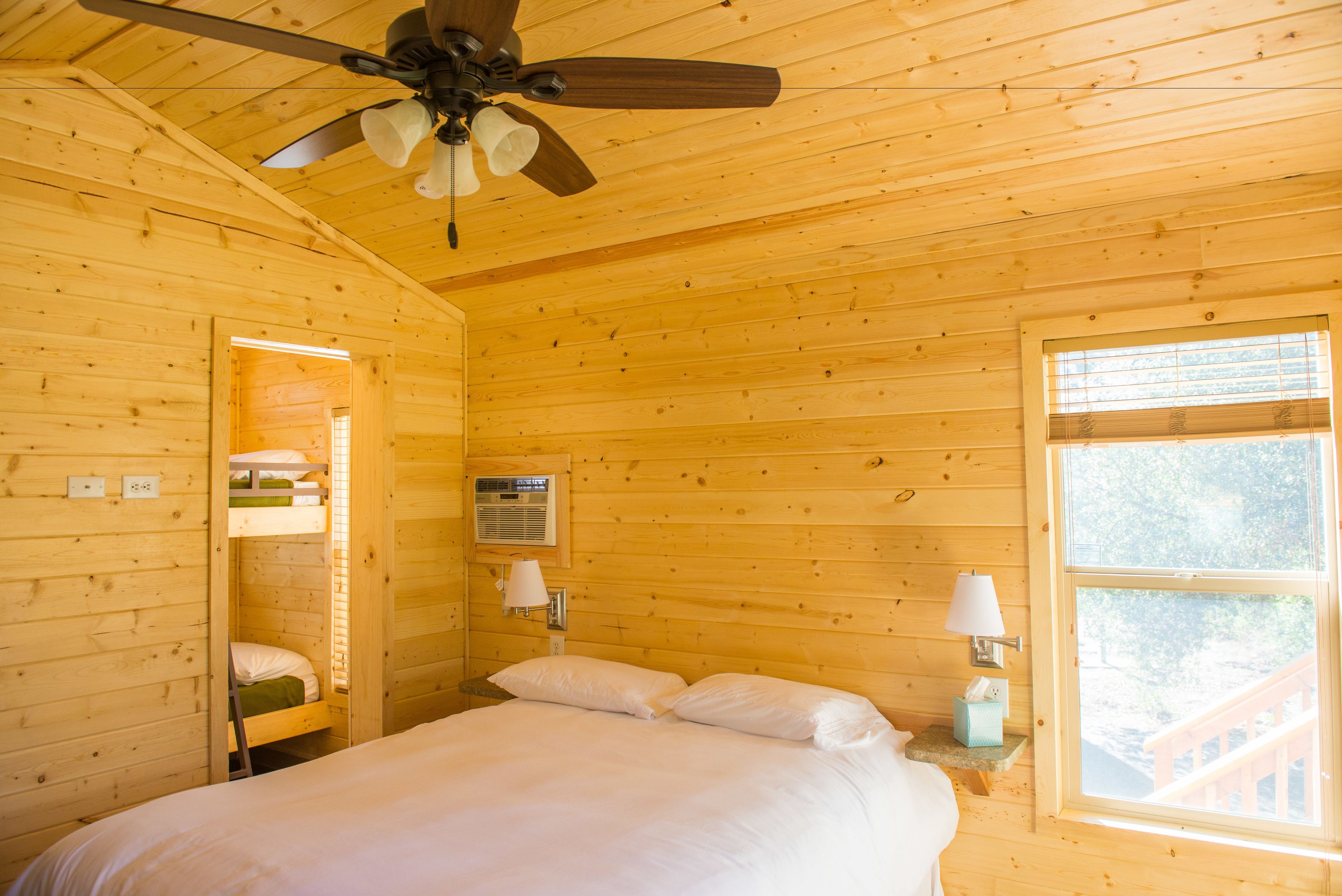 Un dormitorio tipo cabaña con una cama litera, ventilador de techo, ventana con vistas y una lámpara de noche.