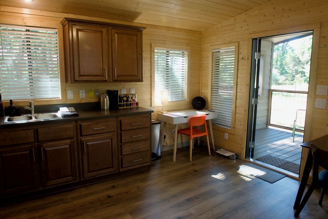 Interior de una cabaña de madera con cocina, una mesa de comedor y una puerta corredera de vidrio que da a una terraza exterior.