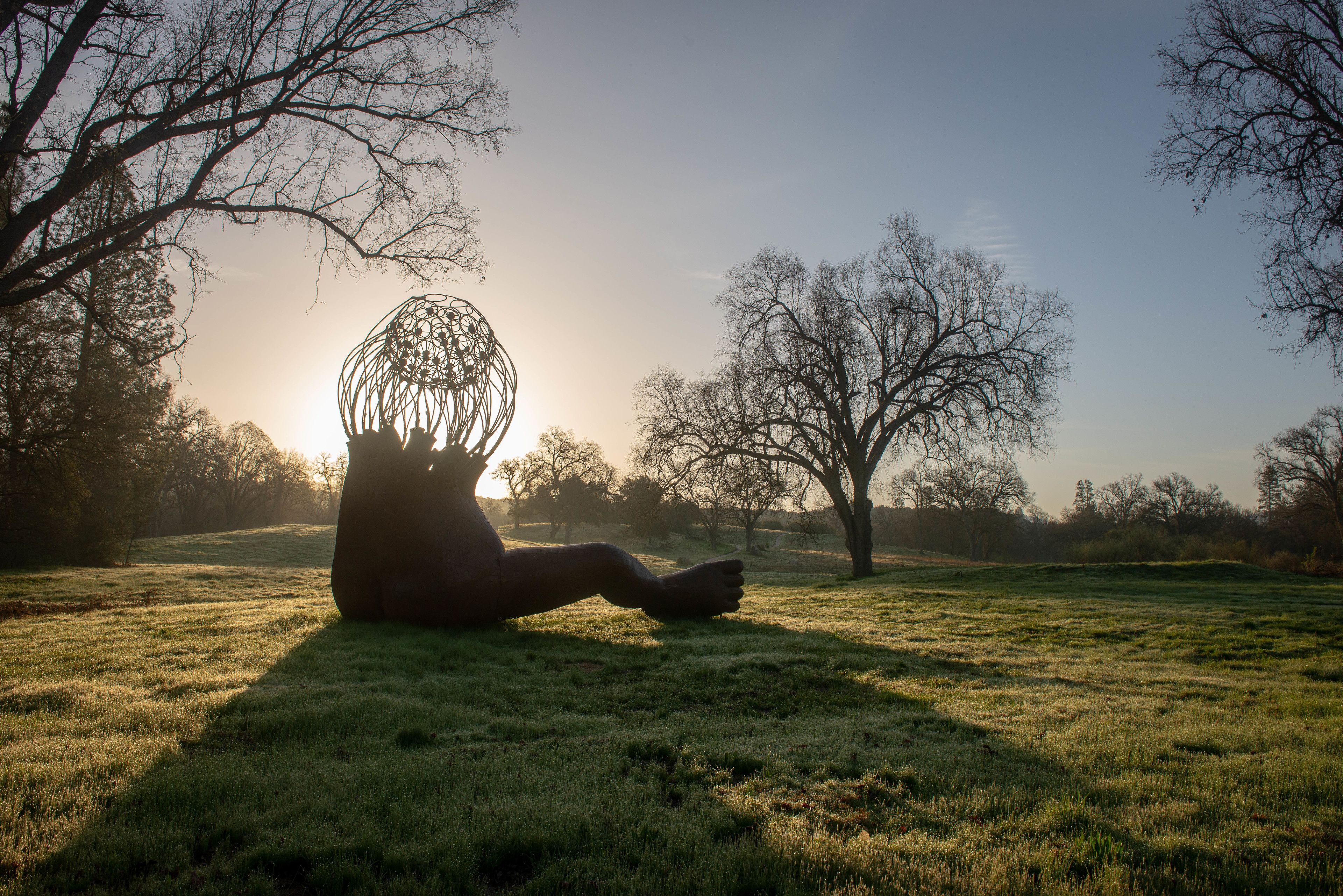 Escultura de una figura humana en un parque con árboles y cielo despejado.