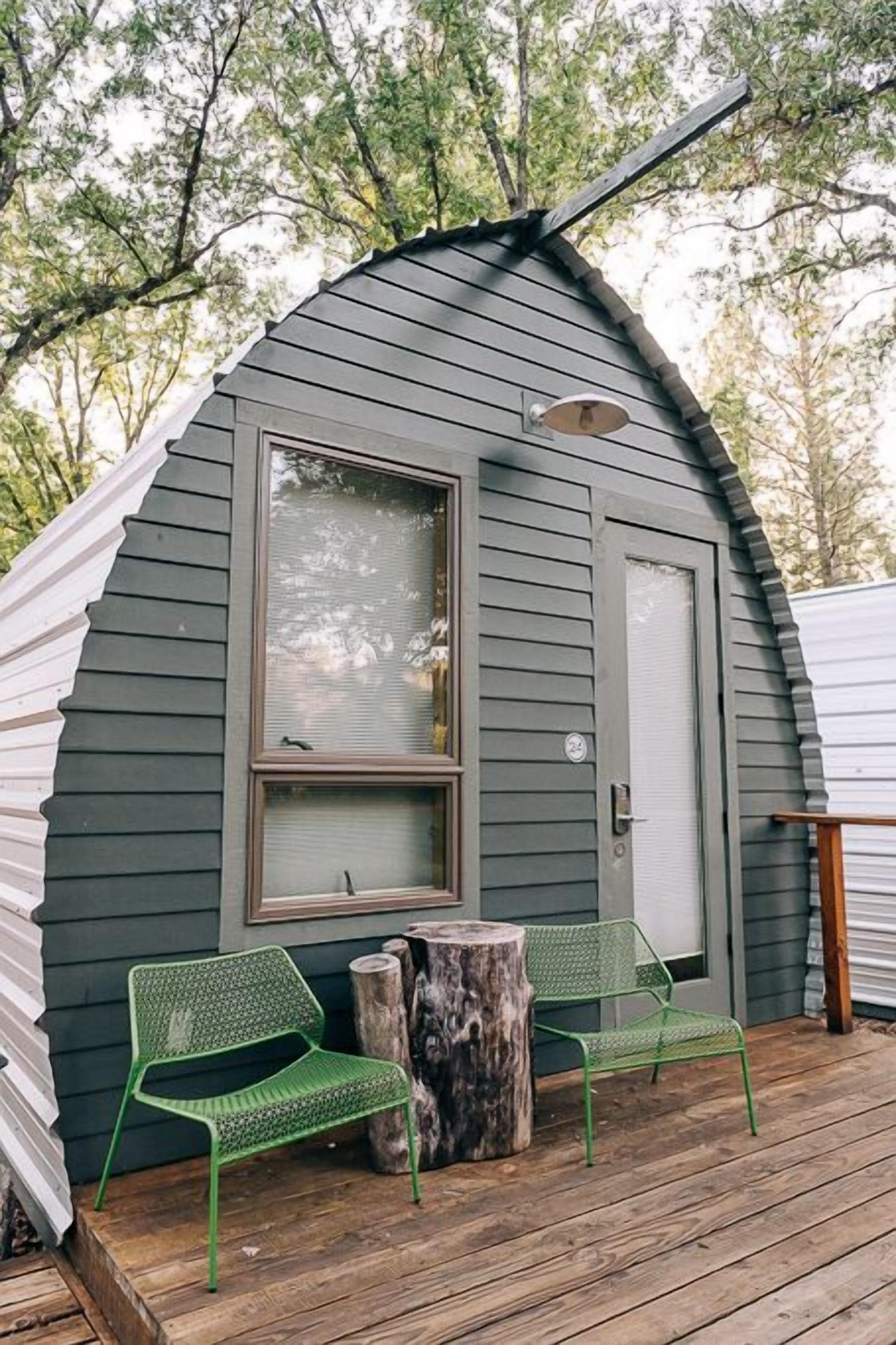 A small, single-story cabin with a sloped roof, a large window, and a door. There are two green metal chairs and a wooden table on the deck.