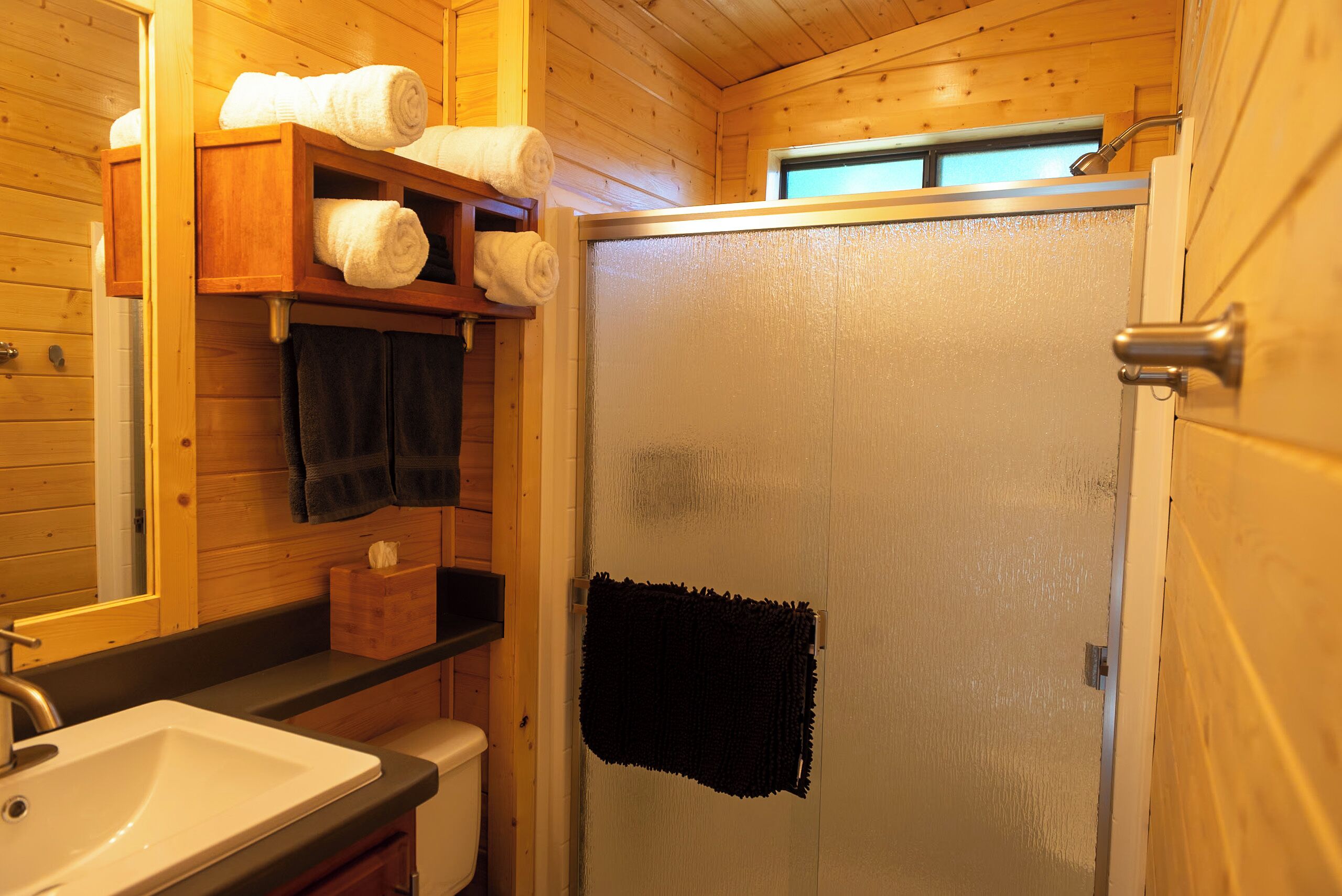 A wooden cabin bathroom with a white sink, a wooden shelf with towels, and a glass shower door.