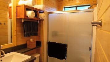 A wooden cabin bathroom with a white sink, a wooden shelf with towels, and a glass shower door.