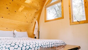 Un dormitorio con techo de madera, cama con ropa de cama estampada, una mesa y dos ventanas con vistas a árboles.