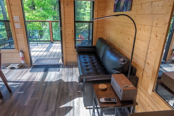 Interior acogedor de una cabaña con un sofá de cuero negro, una mesa de madera con un control remoto y un amplio ventanal con vistas al exterior.