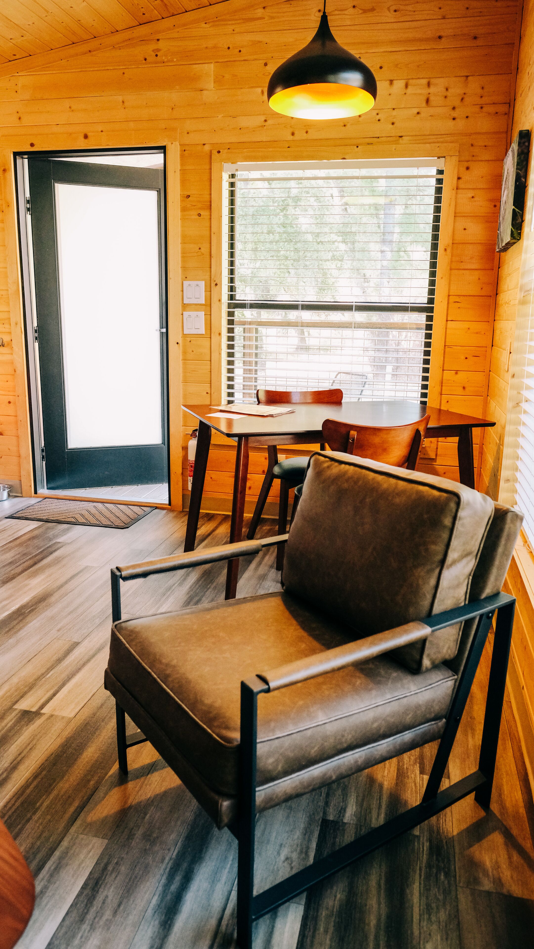 A wooden cabin interior with a dining table, chairs, a sofa, and a large window with blinds.