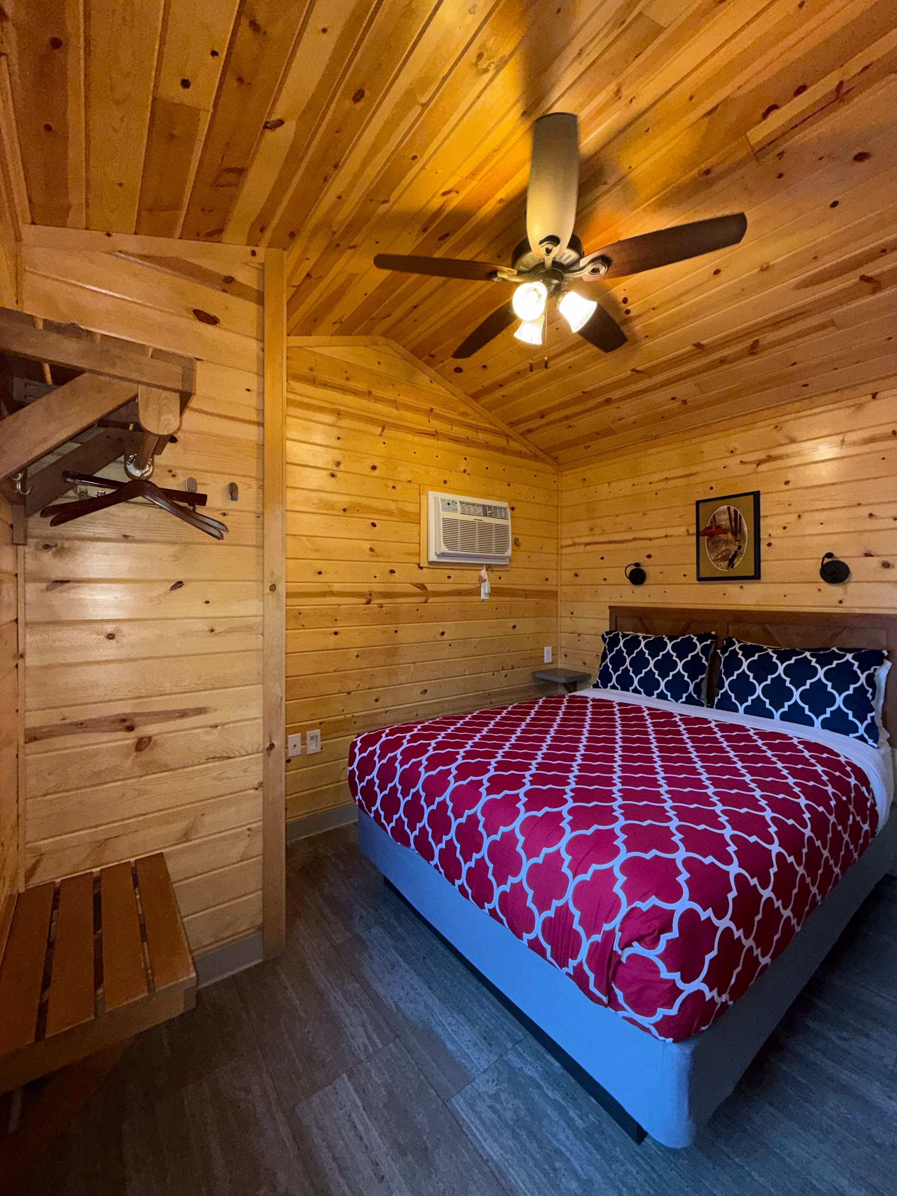 Une chambre dans une cabane en bois, avec un lit, un ventilateur de plafond, un appareil de climatisation et un tableau encadré au mur.