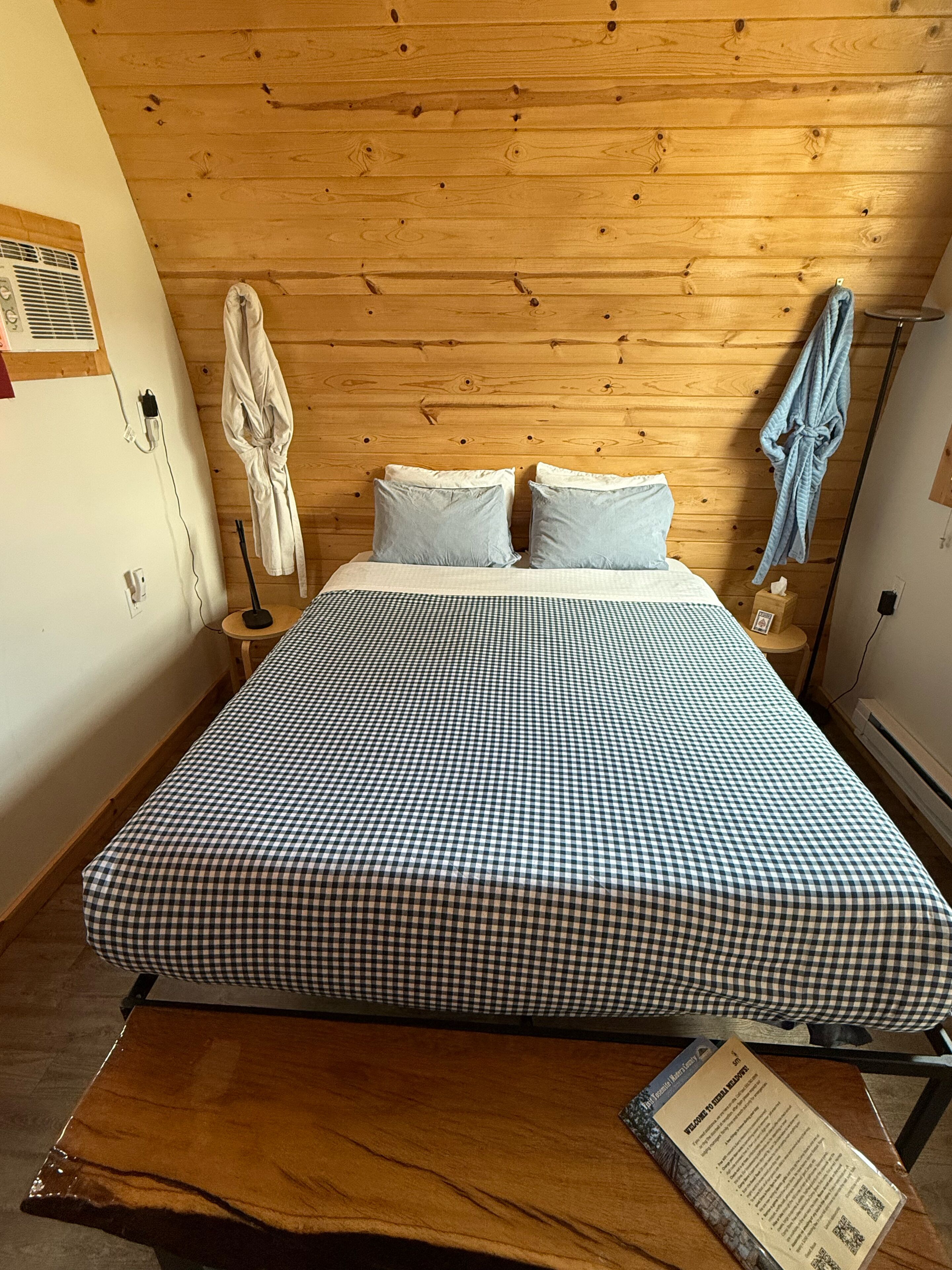 Ein Zimmer mit Holzbalkendecke, einem Bett mit kariertem Überwurf, zwei Kissen, einem an der Wand hängenden Bademantel und einem Nachttisch mit Lampe.