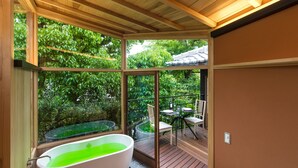Un baño moderno con una bañera independiente, ventanas amplias y vistas a una terraza de madera y follaje exterior.