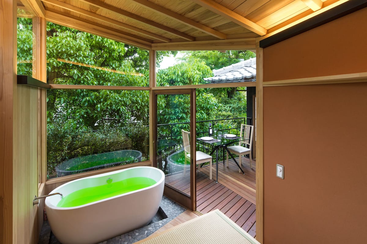 A modern bathroom with a freestanding bathtub, large windows, and a view of a wooden deck and greenery outside.