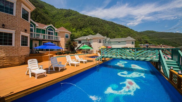 Zona de piscina con sillones de playa y sombrillas, rodeada de edificios con vista a montañas.
