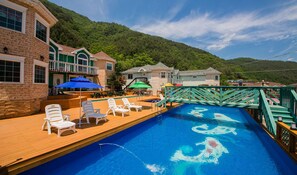 Zona de piscina con sillones de playa y sombrillas, rodeada de edificios con vista a montañas.