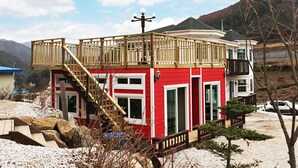 Un edificio de dos pisos rojo y blanco con un balcón, una escalera y un pequeño jardín.